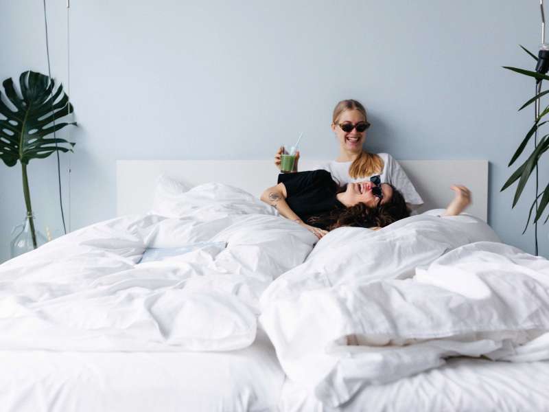 Two friends are lying in bed, drinking and laughing.