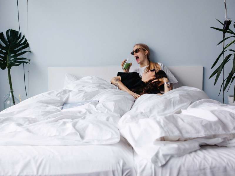 Two friends in bed, one sitting, the other lying down, both laughing.